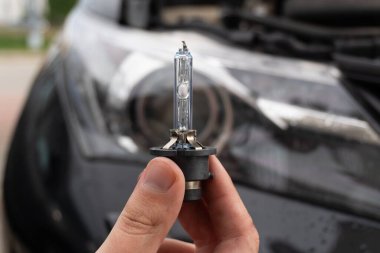 A man's hand holds a single xenon bulb against the blurred background of a car headlight. Replacing the bulb yourself is an important part of ensuring road safety and visibility at night. clipart