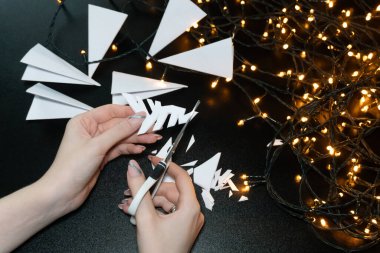 Bir kadın, elinde makasla kar tanelerini keserken, siyah bir arkaplana karşı sıcak sarı bir çelenkle aydınlatılıyor. Parlayan ışıkların ve beyaz kağıdın kontrastı tatil fotoğrafına biraz yaratıcılık ve sihir katıyor..