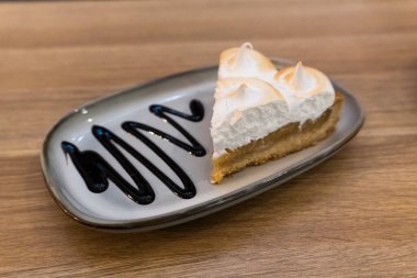 Hassas karamelize kremayla kaplı bir dilim limonlu pasta ahşap bir masanın üzerinde duruyor. Dilim gri bir tabakta duruyor. Yanında dikkatlice serpiştirilmiş sos damlaları var. Tatlının tazeliğini ve canlılığını arttırıyor..