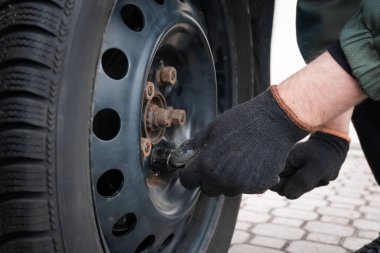 Tekerlek değiştirme işlemi devam ediyor: Siyah eldivenli bir elin çelik bir tekerleğin cıvatalarını hassasiyetle ayarladığı görülüyor. Araç, taşlarla döşenmiş bir yüzeyde, araç güvenliği ve işlevselliğini korumak için atılan rutin bir adımın altını çiziyor.