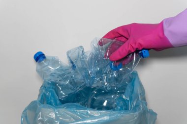 Atık ayıklama: Pembe lastik eldivenli bir kadının eli temizlik sırasında boş bir plastik şişeyi beyaz bir çöp kutusuna koyar. Fotoğraf, çevresel sorumluluğun önemini vurguluyor..