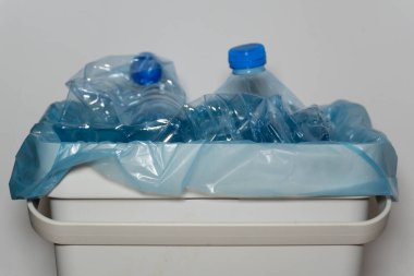 Plastik şişeli çöp tenekesi: Buruşmuş plastik şişelerle dolu mavi plastik torbalı beyaz çöp tenekesi. Görüntü, plastiğin geri dönüşümünün önemini vurguluyor.