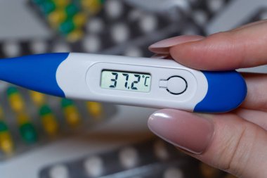 Kullanılan tıbbi ekipman: Mavi uçlu elektronik termometre normal vücut ısısının 37.2C olduğunu gösteriyor. İlaç paketlerinin arka planı iyileşme ve önleme bağlamını vurgulamaktadır.