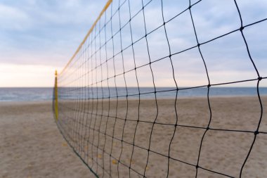 Yumuşak kumların denizle buluştuğu ıssız bir plaja uzanan bir voleybol ağı. Altın gün batımı ve ılık esinti, kıyı tatilinin rahatlatıcı atmosferini vurgular..