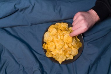 Atıştırmalık: bir adamın eli, tuzlu cips tabağından tek bir cips alır. Sarı çipler ve koyu mavi arkaplan arasındaki zıtlık, çiplerin dokusunu ve çıtırlığını vurgular..