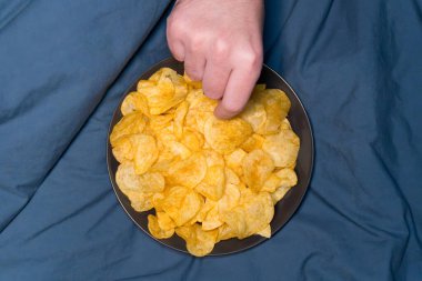 Bir adamın eli dikkatle ince, altın patates cipslerinden bir cips seçer. Yumuşak kumaş ve doğal ışıklandırmayla rahat bir ortam yaratarak bu lezzetli ikramlardan zevk alıyoruz..