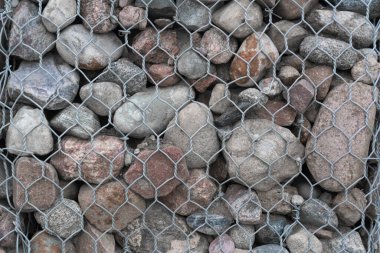 Bir ağ içinde taş çit: Sıkıca paketlenmiş çeşitli boyut ve dokulara sahip bir Gabion 'un yakın çekimi, doğal malzemelerin ve endüstriyel tasarımın kombinasyonunu vurguluyor.