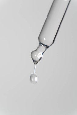 Serum damlası olan pipetin ayrıntıları: Tarafsız beyaz arka planda şeffaf bir pipet düşmeye hazır tek bir damla yüz serumu serumu sergiliyor. Pipetin temiz çizgileri ve parıldayan sıvı yüz serumunun zarafetini vurgular..
