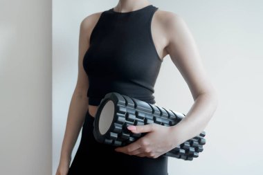 Siyah spor kıyafetli genç bir kızın elindeki masaj silindiri. Spor yaptıktan sonra kas gevşemesi ve iyileşme ekipmanları. Siyah bir spor kıyafeti giyiyor..