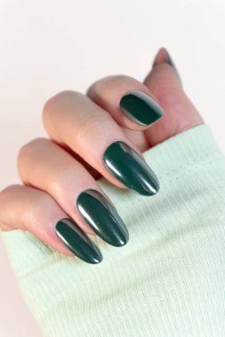 Koyu yeşil cilalı bir avuç içi, stüdyo tarzı bir fotoğrafta koyu renk, parlak ve kusursuz bir şekil kombinasyonu. El kısmen pastel yeşil bir kolla kaplıdır ve tırnak rengiyle uyumlu bir kontrast yaratır..