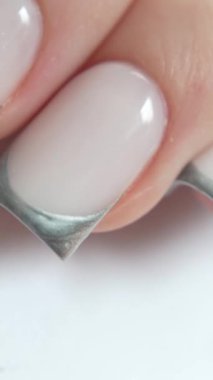 Siyah eldivenli profesyonel manikürcü müşterileri gümüş Fransız manikürcüsüyle tutuyor. Beyaz arkaplan, temiz ve minimal stil. Profesyonel tırnak işçiliğinin hassasiyeti ve özeni vurgulanıyor. Ders ve güzellik eğitimi içeriği için mükemmel