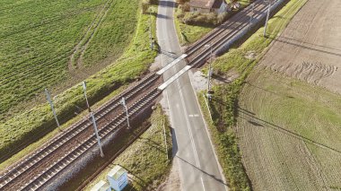 Yeşil ve kahverengi tarlalarla çevrili kırsal bir yoldan geçen bir demiryolu manzarası. Görüntü, ulaşımın ve kırsal kesişimlerin sakin ve manzaralı ortamlarını yakalıyor..