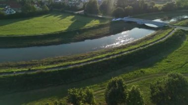 Hava fotoğrafı, yeşil tarlalar ve yumuşak güneş ışığında gölgeli ağaçlarla çevrili zarif bir köprünün geçtiği bir nehri gösteriyor..