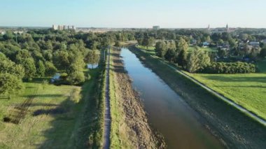 Bu hava fotoğrafı, yemyeşil tarlalarla çevrili dolambaçlı bir nehri ve arka planda açık mavi bir gökyüzünün altında uzak bir kasabayı yakalar..