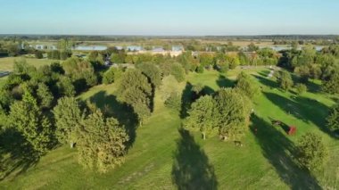 Bu hava görüntüsü, sakin bir nehirle çevrelenmiş çimlerin üzerine uzun gölgeler düşen sayısız ağacın bulunduğu yemyeşil bir park sergiliyor..