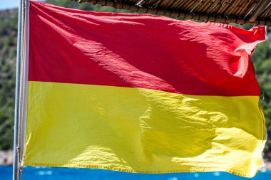 Kumlu bir sahilde rüzgarda dalgalanan cankurtaran bayrağının yakın plan çekimi, canlı deniz manzarası ile güvenlik ve tetikte olmayı sembolize ediyor..