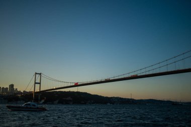 İstanbul Boğazı Köprüsü 'nün gün batımında nefes kesen manzarası, köprünün silueti gökyüzünün canlı renklerine karşı, aşağıdaki sakin sulara yansıyor..