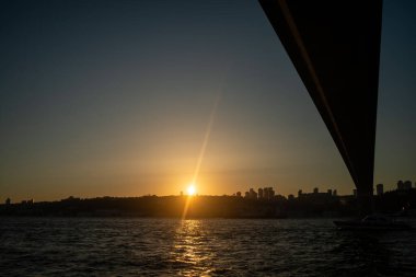 İstanbul Boğazı Köprüsü 'nün gün batımında nefes kesen manzarası, köprünün silueti gökyüzünün canlı renklerine karşı, aşağıdaki sakin sulara yansıyor..