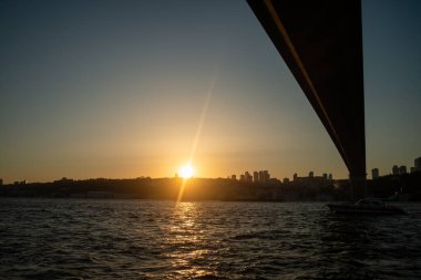 İstanbul Boğazı Köprüsü 'nün gün batımında nefes kesen manzarası, köprünün silueti gökyüzünün canlı renklerine karşı, aşağıdaki sakin sulara yansıyor..