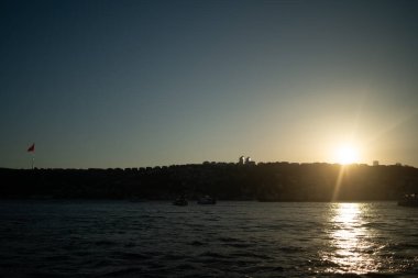 İkonik Türk bayrağının gururla dalgalandığı gün batımında İstanbul Boğazı 'nın nefes kesici manzarası. Altın ışık suyu yansıtıyor, şehrin tarihi siluetini vurguluyor ve sakin, resimsel bir manzara yaratıyor..