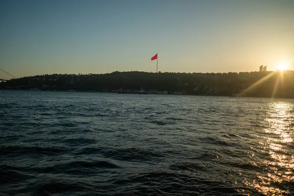 İkonik Türk bayrağının gururla dalgalandığı gün batımında İstanbul Boğazı 'nın nefes kesici manzarası. Altın ışık suyu yansıtıyor, şehrin tarihi siluetini vurguluyor ve sakin, resimsel bir manzara yaratıyor..