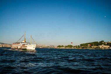 İstanbul 'daki Boğaz' ın pitoresk bir manzarası, arka planda şehrin ufuk çizgisi ve ikonik simgeleriyle birlikte suda yüzen bir yolcu feribotu yer alıyor..