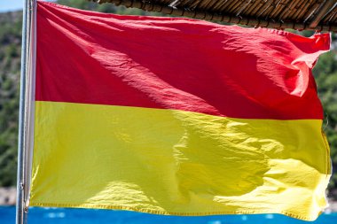 Kumlu bir sahilde rüzgarda dalgalanan cankurtaran bayrağının yakın plan çekimi, canlı deniz manzarası ile güvenlik ve tetikte olmayı sembolize ediyor..