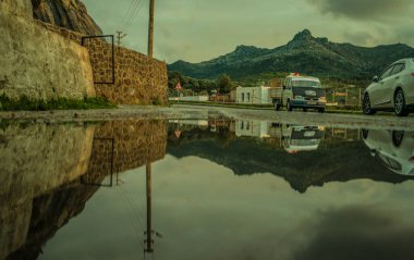 Yağmurlu köyde, yağmurlu havanın dingin güzelliğinde yakalanan ıslak yol arabaları, dağlar ve köy hayatı üzerine yansıtıcı bir sahne gözler önüne seriliyor.