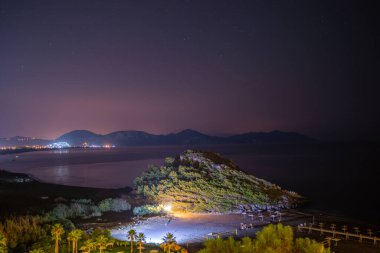 Yıldızlarla dolu bir gökyüzünün altında denizin dingin güzelliğini gösteren nefes kesici uzun pozlu bir fotoğraf. Ufuk çizgisine hafifçe çizilmiş uzak bir ada..