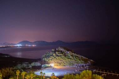 Yıldızlarla dolu bir gökyüzünün altında denizin dingin güzelliğini gösteren nefes kesici uzun pozlu bir fotoğraf. Ufuk çizgisine hafifçe çizilmiş uzak bir ada..