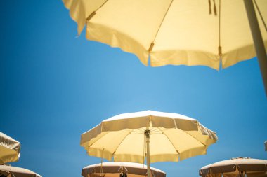 Aşağıdan görülen canlı bir plaj şemsiyesi, açık mavi gökyüzü ve arka planda altın güneş ışığı.