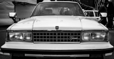 A vintage car captured in a black and white photo, showcasing its distinct boxy design and prominent headlights. Parked at an outdoor event, the car is surrounded by tents and other temporary structures, suggesting a car show atmosphere. clipart