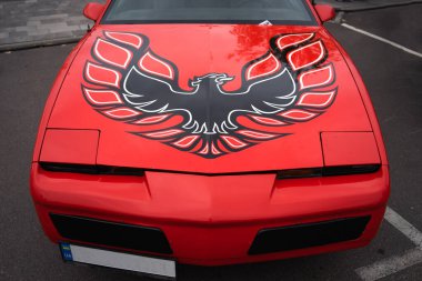 Kaputu siyah ve kırmızı bir Anka Kuşu desenli, göz alıcı kırmızı bir araba park halinde. Otomotiv meraklıları ve özel otomotiv meraklıları için, çıkartmanın çarpıcı tasarımı arabaya ayırt edici ve çarpıcı bir özellik kazandırıyor..