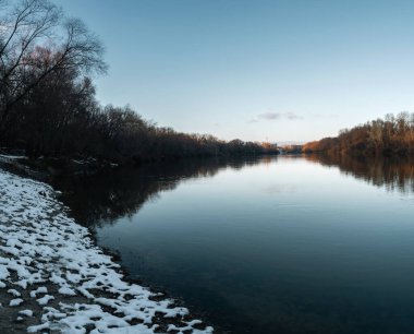Gün batımında kış ağaçlarını yansıtan sakin bir nehri olan karla kaplı nehir kıyısı..