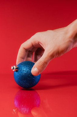 Yansıtıcı kırmızı bir yüzey üzerinde mavi bir Noel süslemesi tutan bir el ile neşeli bir sahne.
