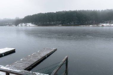 Sisli bir gökyüzünün altında sakin, buzlu bir kış gölüne uzanan ahşap bir iskele..