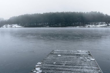 Sisli bir gökyüzünün altında sakin, buzlu bir kış gölüne uzanan ahşap bir iskele..