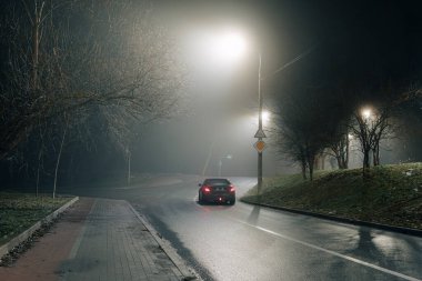 Islak yolda giden bir arabanın olduğu sisli şehir gece sahnesi. Çıplak ağaçlarla çevrili, parlayan sokak lambalarıyla aydınlatılmış..