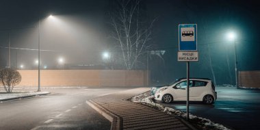 Gece sisli bir şehir otoparkında otobüs durağı tabelasının yanına park edilmiş, sokak lambalarıyla aydınlatılmış küçük beyaz bir araba..