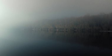 Gece sisli bir nehir kenarında, uzak ışıklarla aydınlatılan sakin bir su yüzeyi ve karanlık, sakin bir kıyı şeridi var..