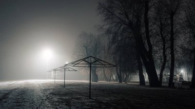 Gece sisli bir nehir kenarında, uzak ışıklarla aydınlatılan sakin bir su yüzeyi ve karanlık, sakin bir kıyı şeridi var..