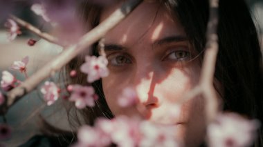 Kiraz çiçekleriyle çerçevelenmiş bir kadının yumuşak doğal portresi. Hassas ışıklandırma ve sığ alan derinliği ruhani bir bahar havası yaratır..