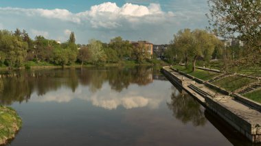 Sakin suyu, ağaçları, bulutları ve mavi gökyüzünü yansıtan manzaralı bir nehir manzarası. Taş seti ve yeşillik, doğa, seyahat ve açık hava rahatlama temaları için mükemmel, huzurlu bir atmosfer yaratır..