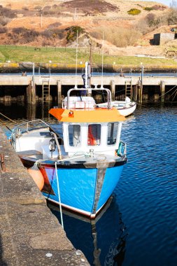 A blue and white fishing boat is moored against a harbour wall. In the middle distance, there is a jetty, running along the length of the image and above that, there is a grassy hillside. clipart