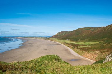 Gower Yarımadası 'ndaki Rhossili Sahili' ne tepeden bakarken mavi gökyüzünün altında renkli tepeleri olan büyük, ıssız kumlu bir sahil görünüyor.