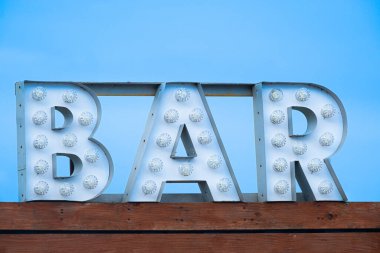A cut-out metal bar sign with light bulbs sits atop a building, photographed in daytime against a clear blue sky