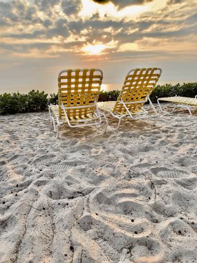 Two white woven chairs with brown rubber straps sit on a pristine sandy beach, facing the ocean during sunset, creating an inviting scene for two to share a tranquil moment while enjoying the beauty of the setting sun over the sea clipart