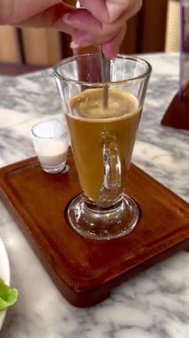 Bir kadının eli, eski tarz kahveyi, tahtadan bir tepside, mermer bir masada süt bardağıyla karıştırır. Sahne, kahve sevenler ve yaşam tarzı tasvirleri için mükemmel bir sıcaklık ve rahatlama sağlar.