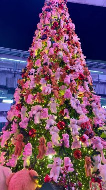 A beautifully lit Christmas tree features sparkling red balls and an assortment of donated teddy bears, predominantly in pink and brown hues. Ideal for use in holiday cards, festive decorations, or social media to convey warmth and happiness. clipart