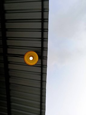 Galvanize çelik çatının altında asılı bir lamba ampulü. Arka plan da bulutlu bir gökyüzü.